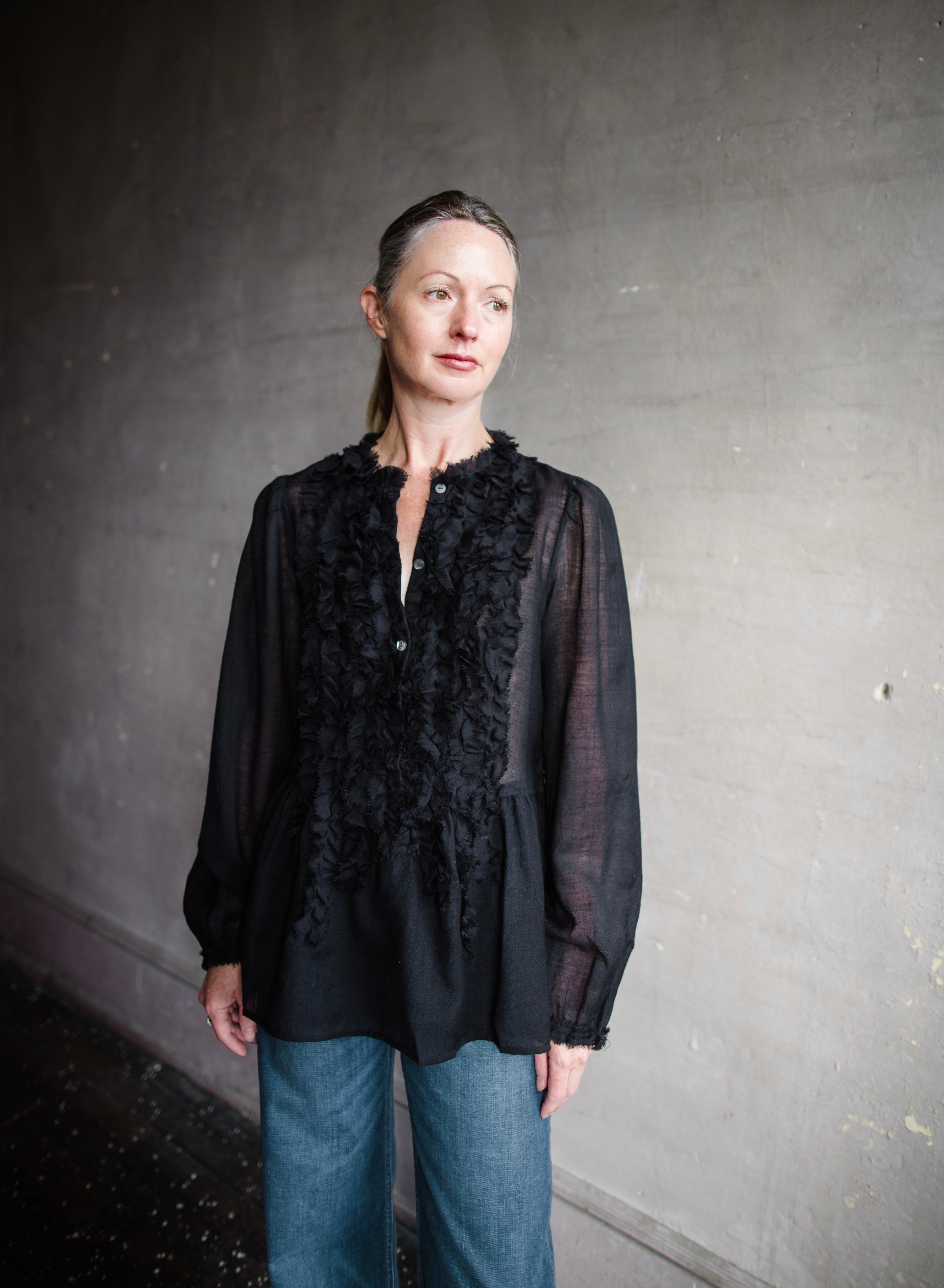 Image features woman wearing The Cosima Tunic by Ulla Johnson in Noir features a button down v neckline with rows of ruffles and iridescent sea shell buttons.