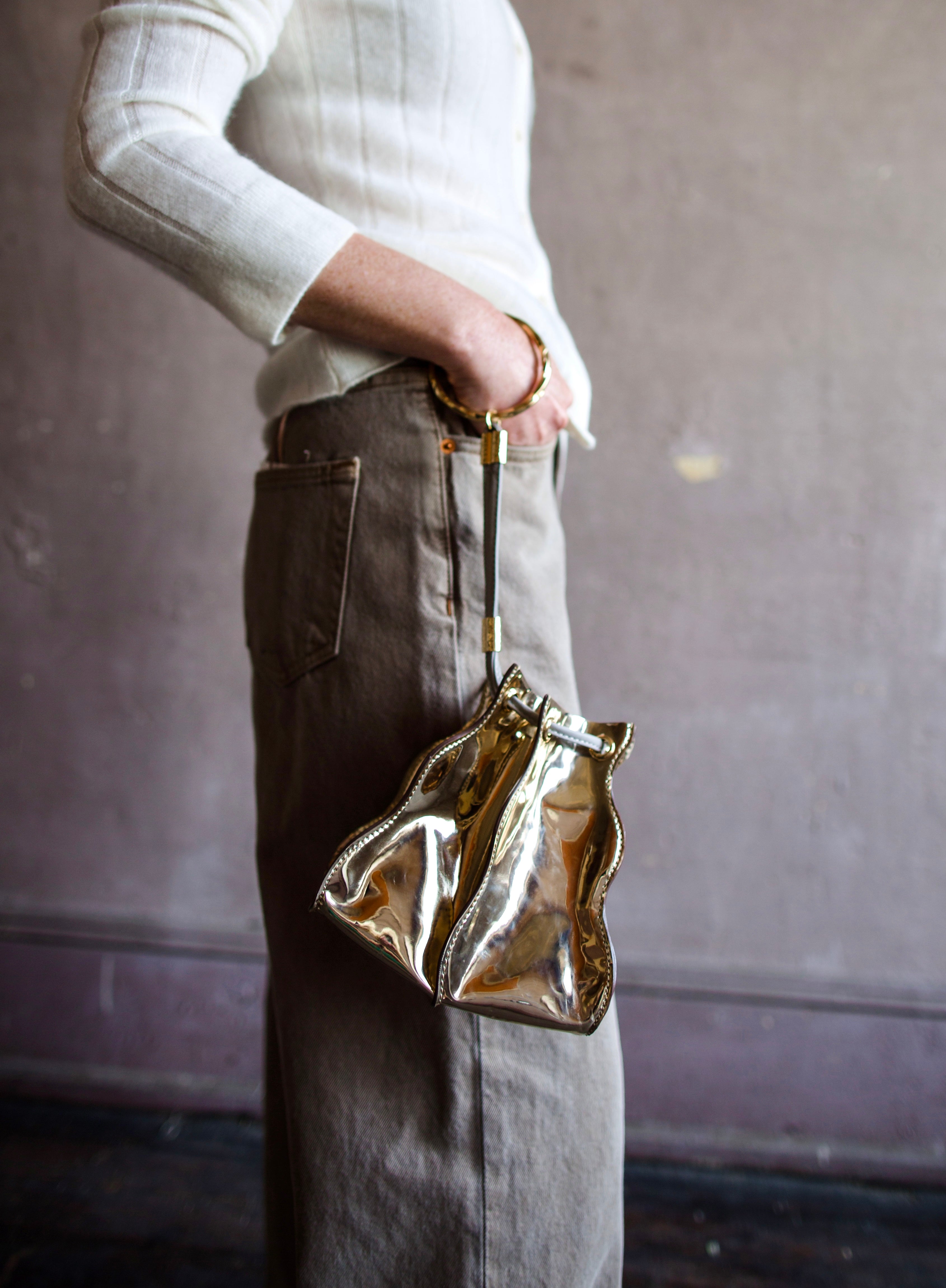 Image featuring a woman wearing the Adria wave pochette by Ulla Johnson with a drawstring closure, a crossbody strap and metal bangle in Platino gold.