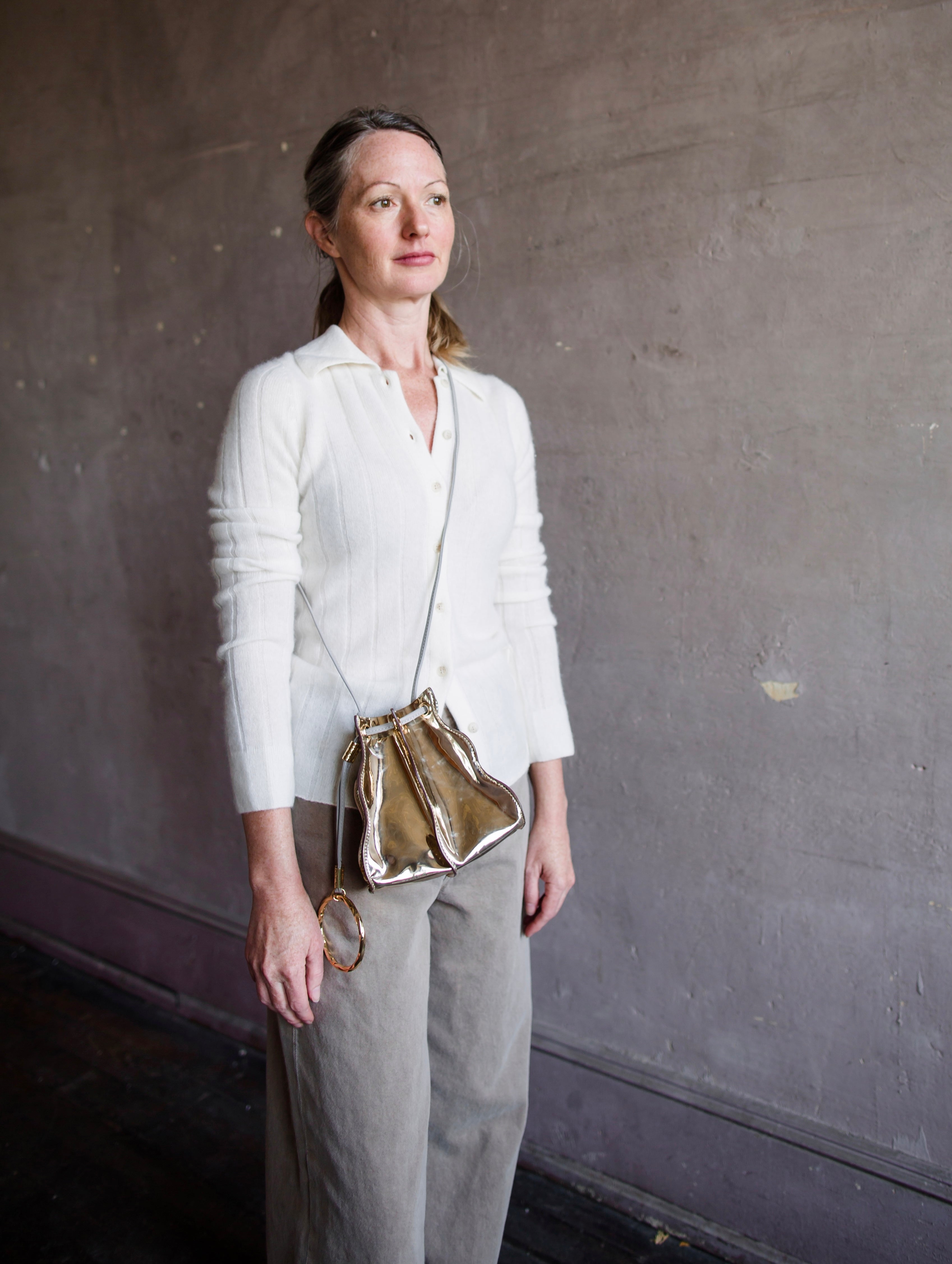 Image featuring a woman wearing the Adria wave pochette by Ulla Johnson with a drawstring closure, a crossbody strap and metal bangle in Platino gold.