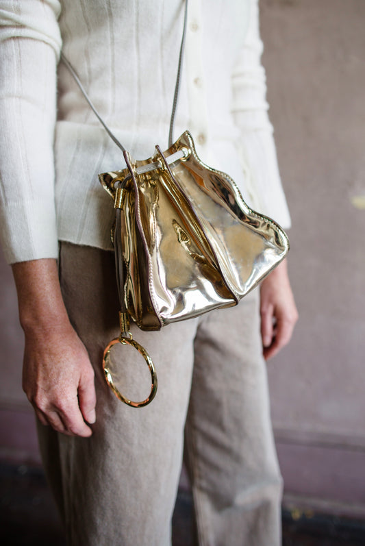 Image featuring a woman wearing the Adria wave pochette by Ulla Johnson with a drawstring closure, a crossbody strap and metal bangle in Platino gold.