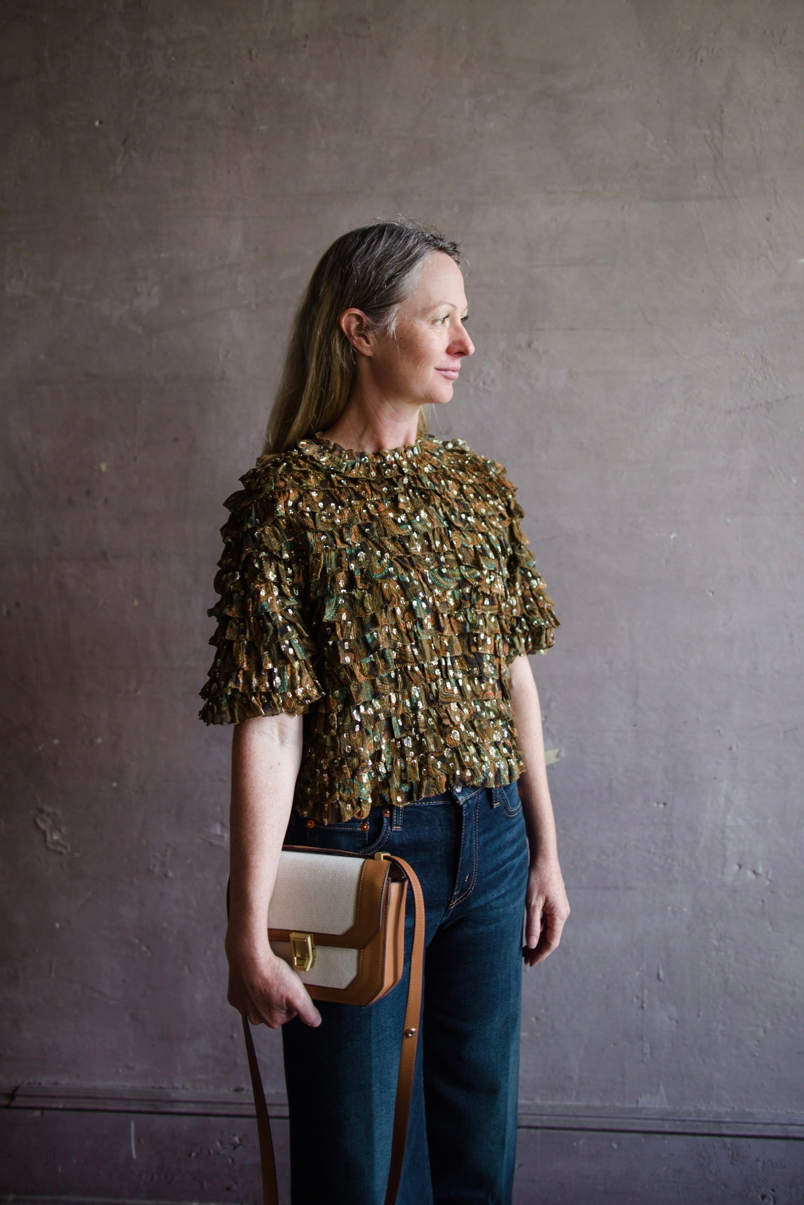 Image featuring a woman wearing the Carmella crinkle silk chiffon top by Ulla Johnson with a crew neckline with tiered ruffles and golden specks throughout.