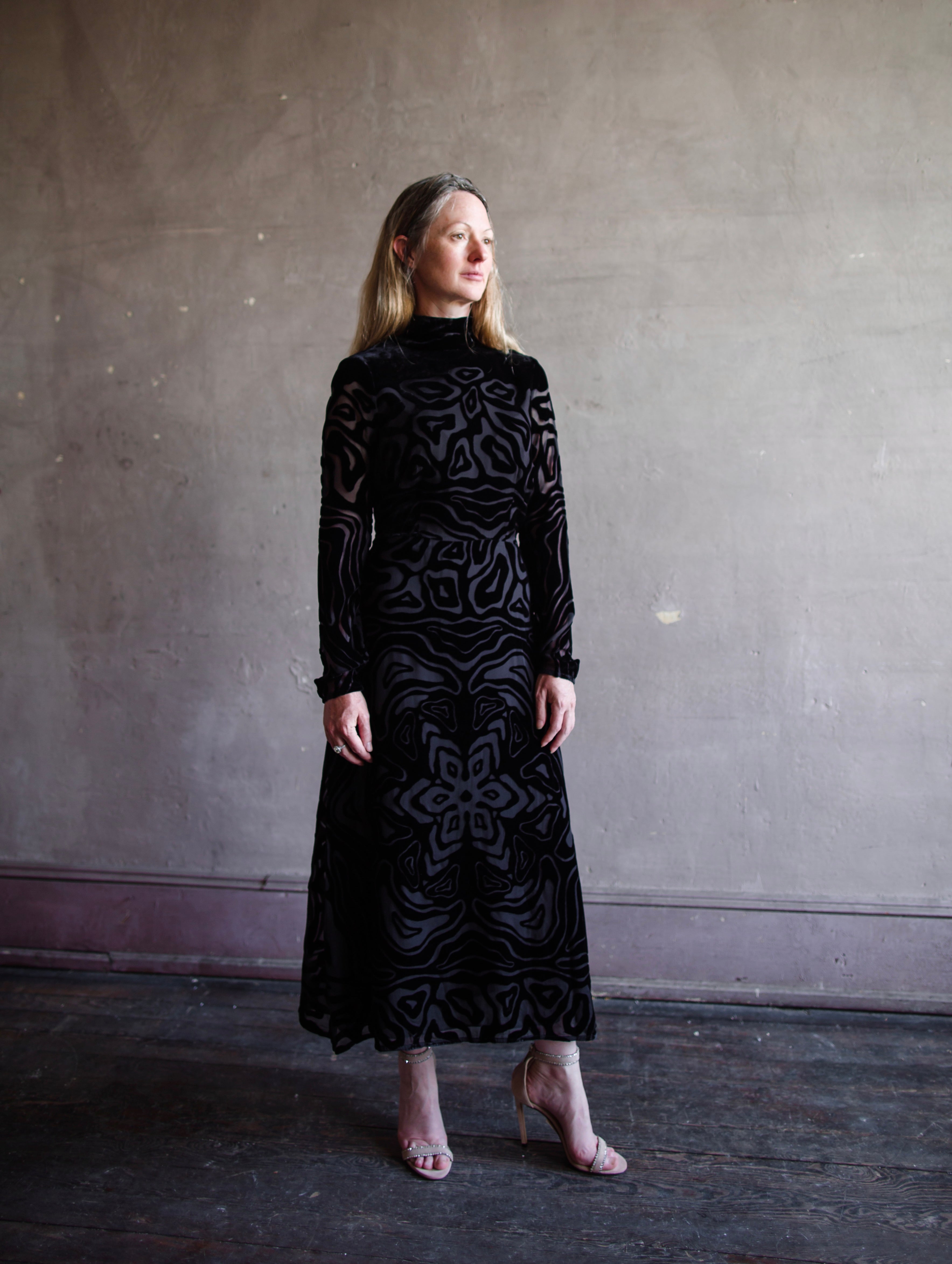 Image featuring a woman wearing the Helena dress by Ulla Johnson with a high neckline, a wavy floral  burnout pattern and maxi hemline in black.