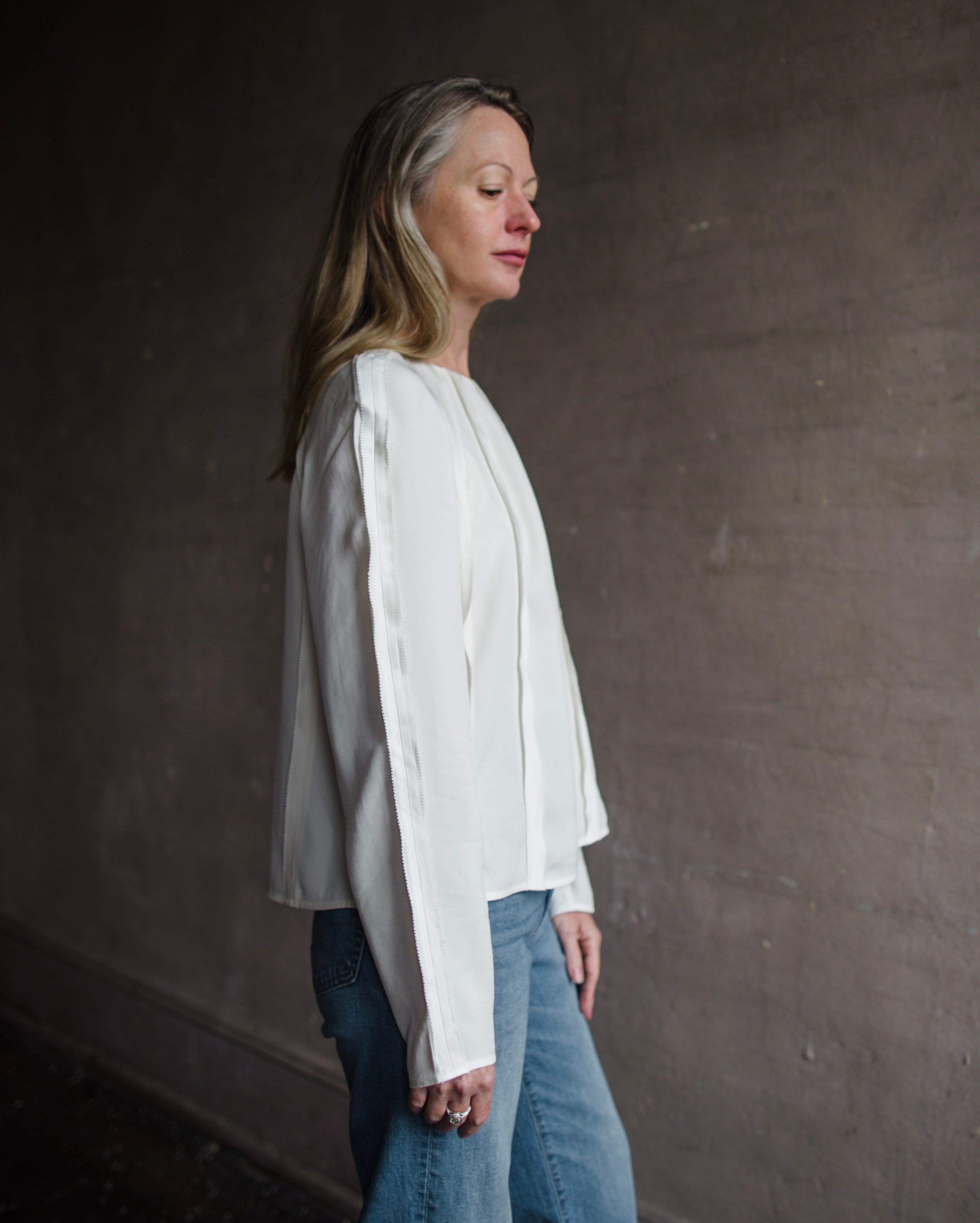 Side profile of mature woman wearing Ulla Johnson Kavi long sleeve Blouse with three dimensional linear seam details crew neck in cream