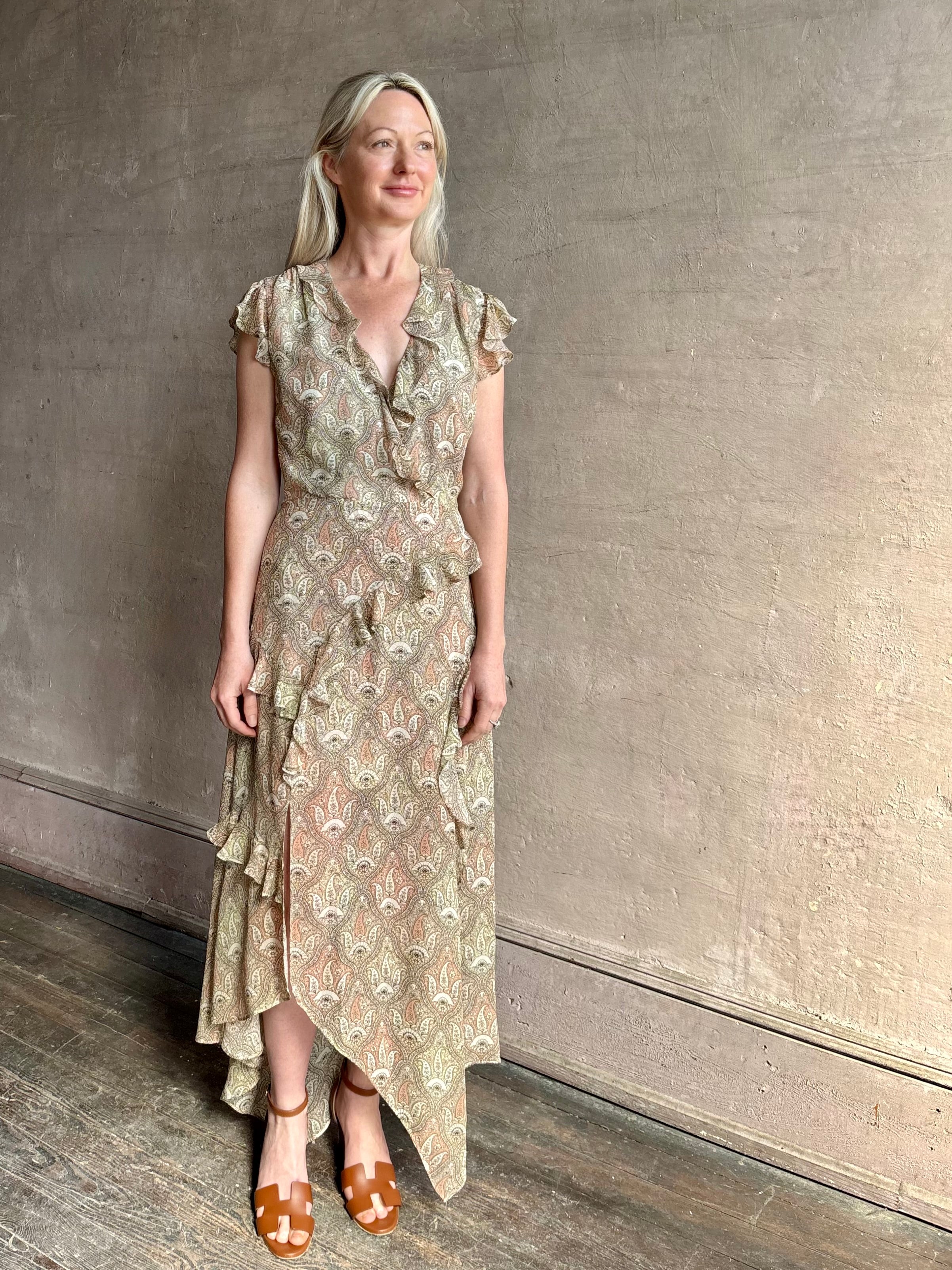 Mature woman wearing the Cap Sleeve Anenel maxi Dress from luxury designer Veronica Beard rendered in a soft khaki, green, and pink paisley print and crafted in silk with cap sleeves, v- neckline, and asymmetrical hem all finished with soft feminine ruffles in a timeless A-line silhouette