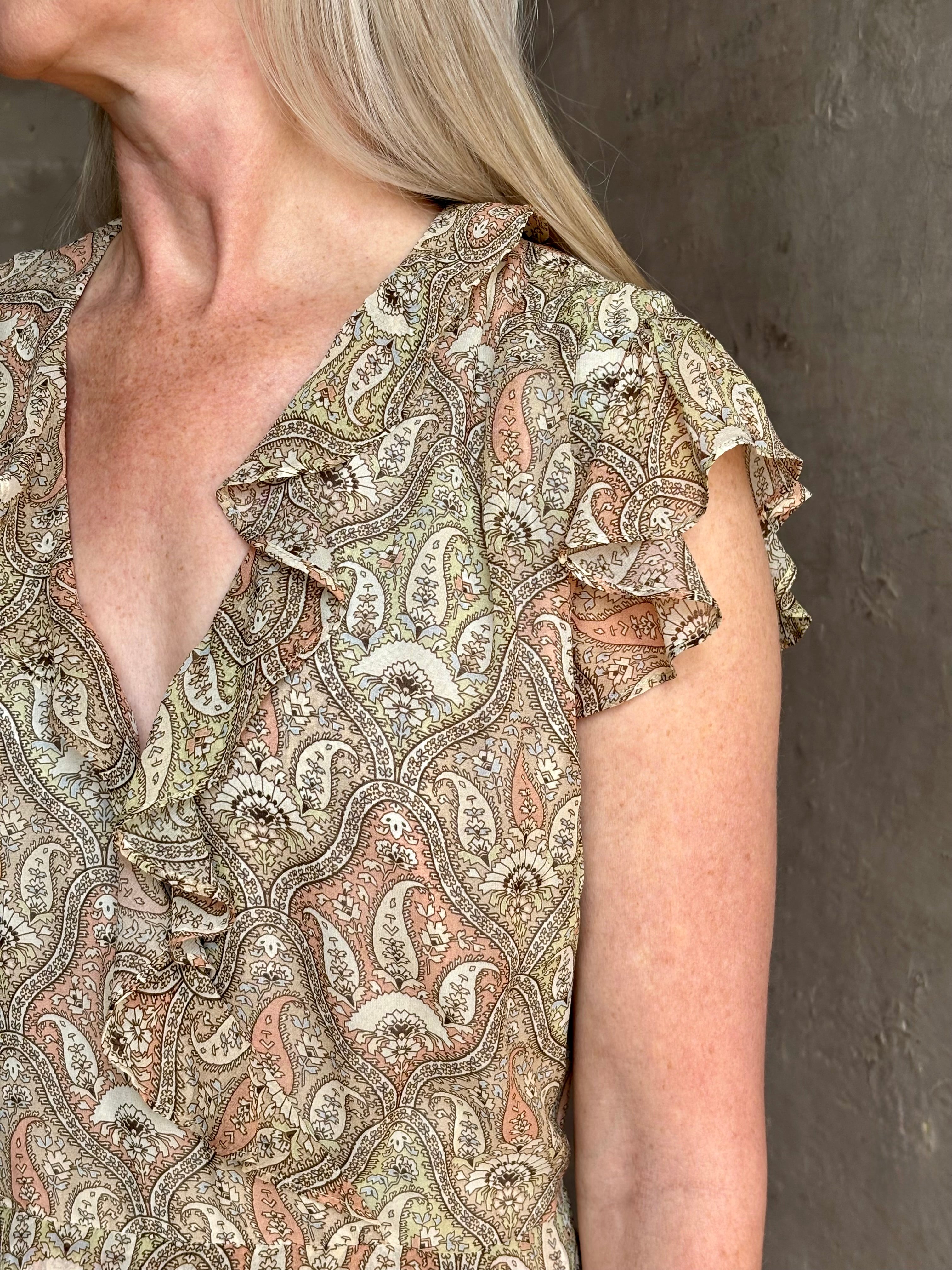 Mature woman wearing the Cap Sleeve Anenel maxi Dress from luxury designer Veronica Beard rendered in a soft khaki, green, and pink paisley print and crafted in silk with cap sleeves, v- neckline, and asymmetrical hem all finished with soft feminine ruffles in a timeless A-line silhouette