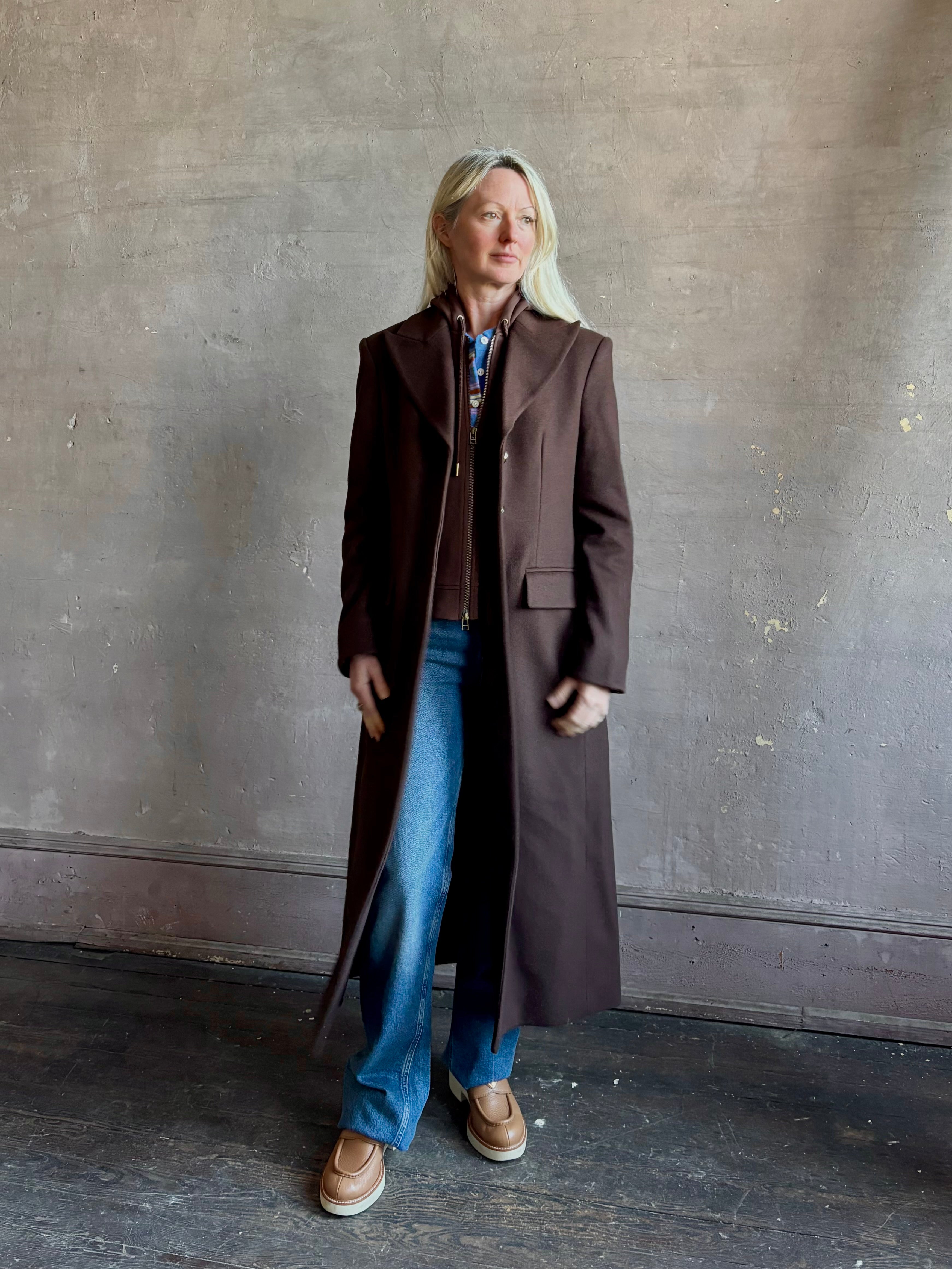Mature woman wearing Veronica Beard Elspeth Coat with OVS Hoodie Dickey in Espresso brown wool-cashmere A-line coat with a removable hoodie layer. Playful, sculptural cold-weather luxury.