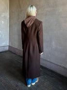 Mature woman wearing Veronica Beard Elspeth Coat with OVS Hoodie Dickey in Espresso brown wool-cashmere A-line coat with a removable hoodie layer. Playful, sculptural cold-weather luxury.