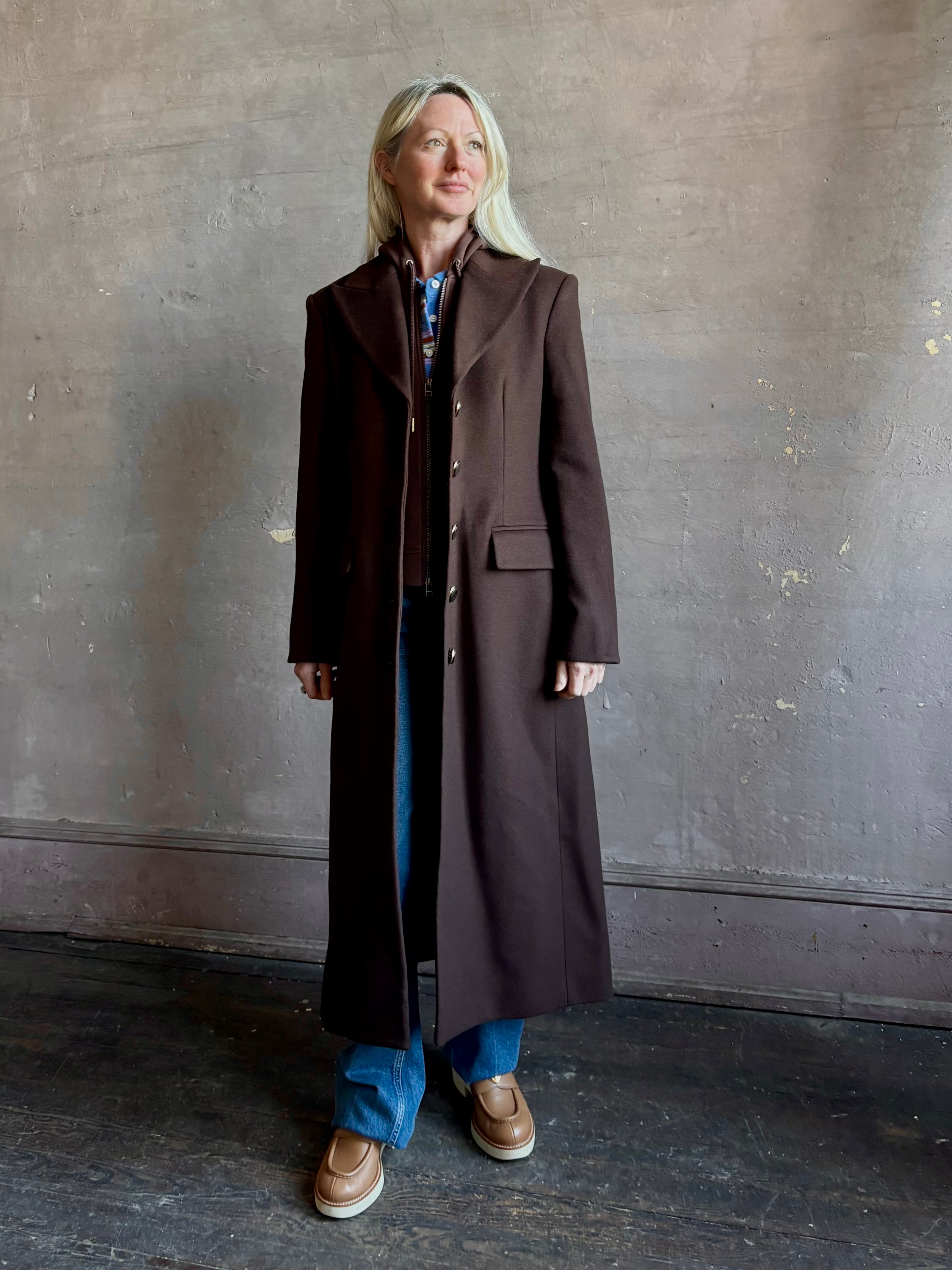 Mature woman wearing Veronica Beard Elspeth Coat with OVS Hoodie Dickey in Espresso brown wool-cashmere A-line coat with a removable hoodie layer. Playful, sculptural cold-weather luxury.