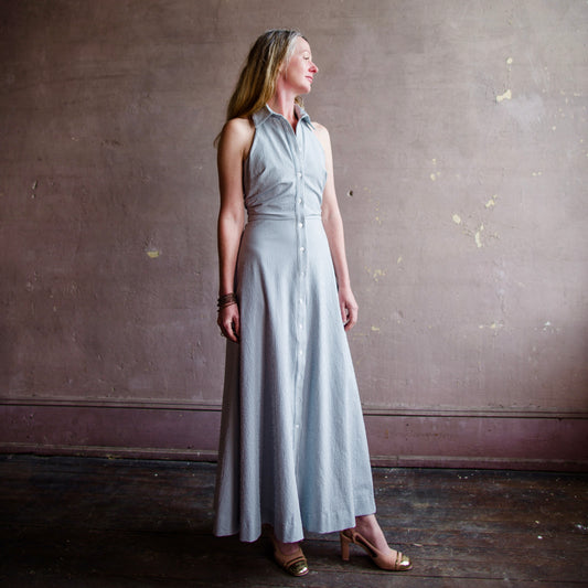 Image featuring a woman wearing the Mackey A-line shirtdress with a halter neckline and backless in a blue and white searsucker fabric.