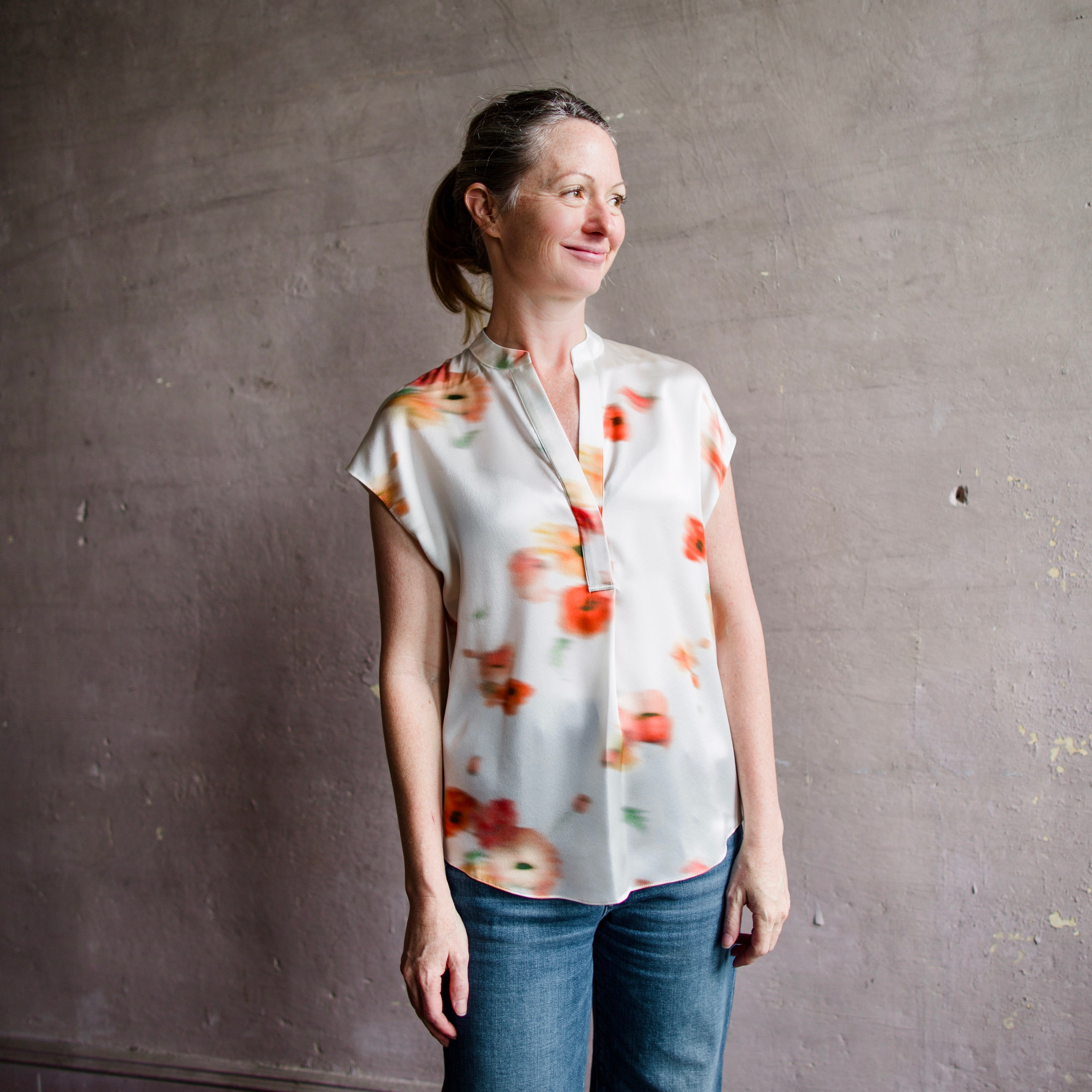 Image featuring a woman wearing the Vince poppy silk blouse with a band collar, split neckline, and short dolman sleeves, and showcases an abstract blurred poppy print.