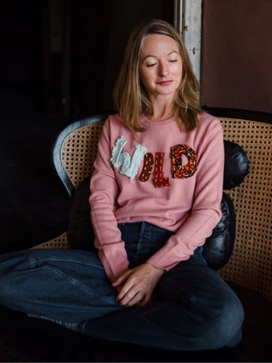 Image featuring a woman wearing the pink cashmere sweater by Lingua Franca with a crew neckline, long sleeves, and hand-applied patchwork lettering "wild" on front.