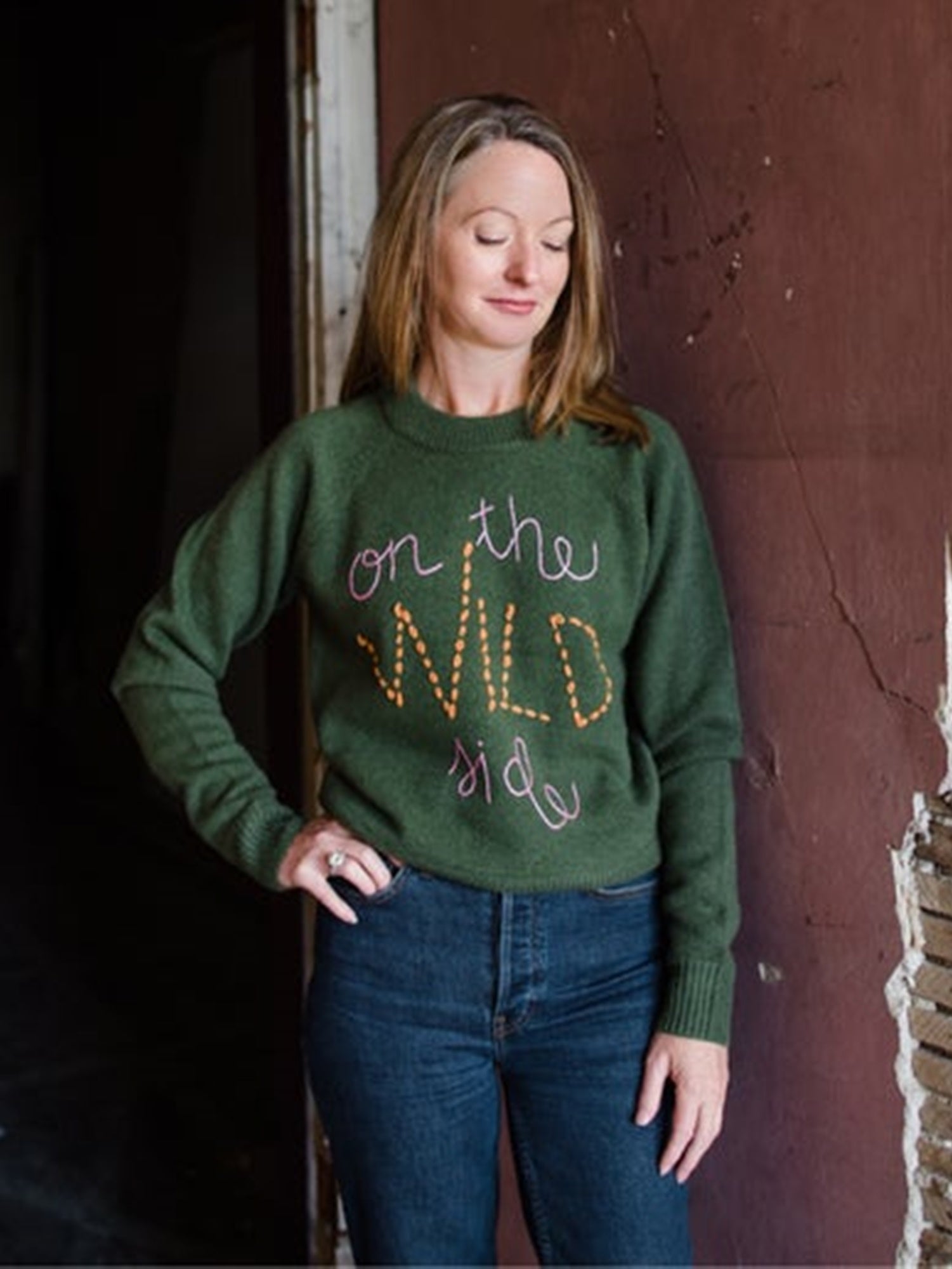 Image featuring a woman wearing the green cashmere and wool sweater by Lingua Franca with a crew neckline, relaxed fit and "on the wild side" embroidered on the front in pink and yellow.