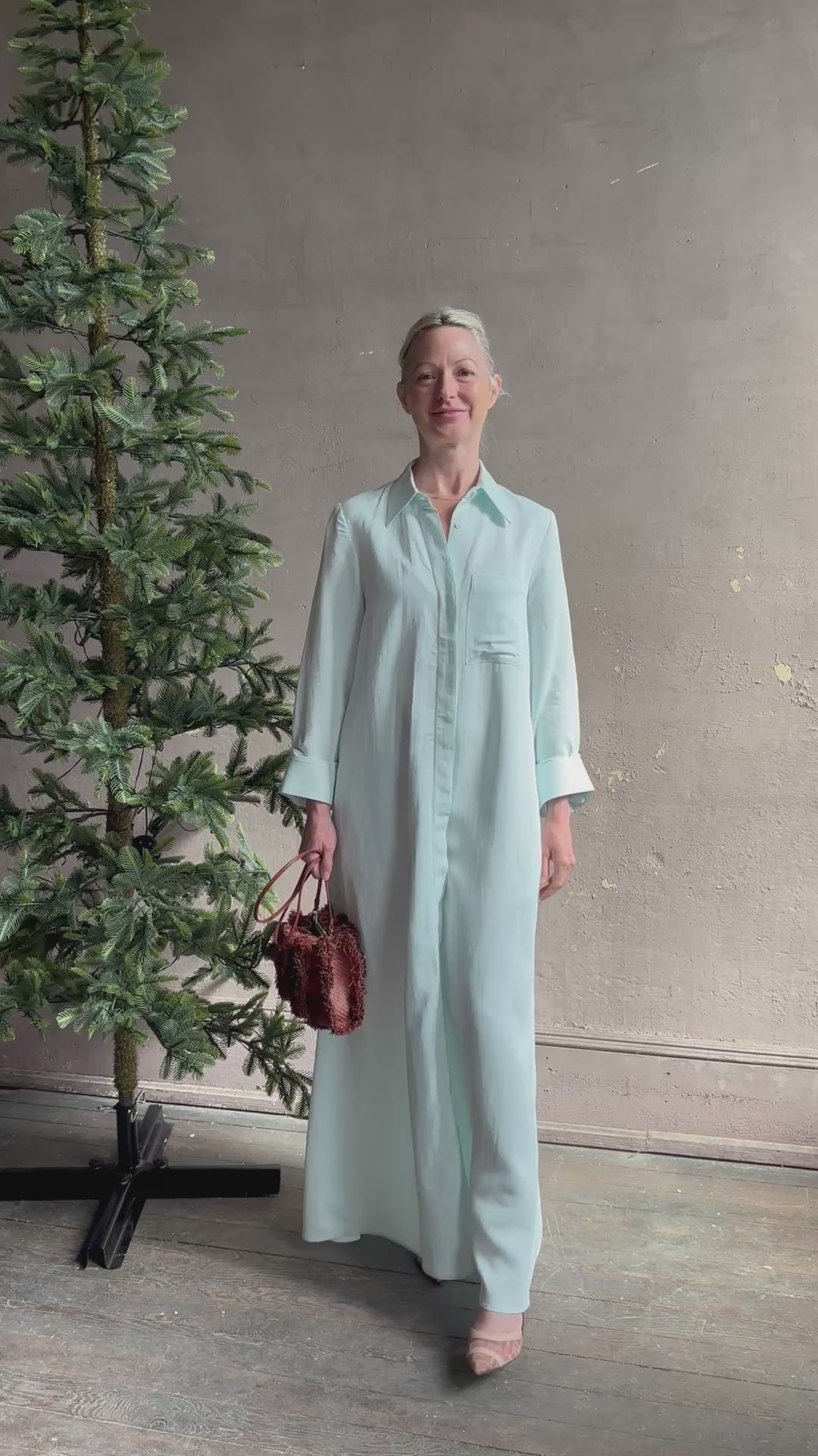 Mature woman reviewing floor-length Jenny's Gown from TWP rendered in a sea glass hue and crafted from a viscose linen blend with beautiful draping, long sleeves, button front and cool girl loose fit