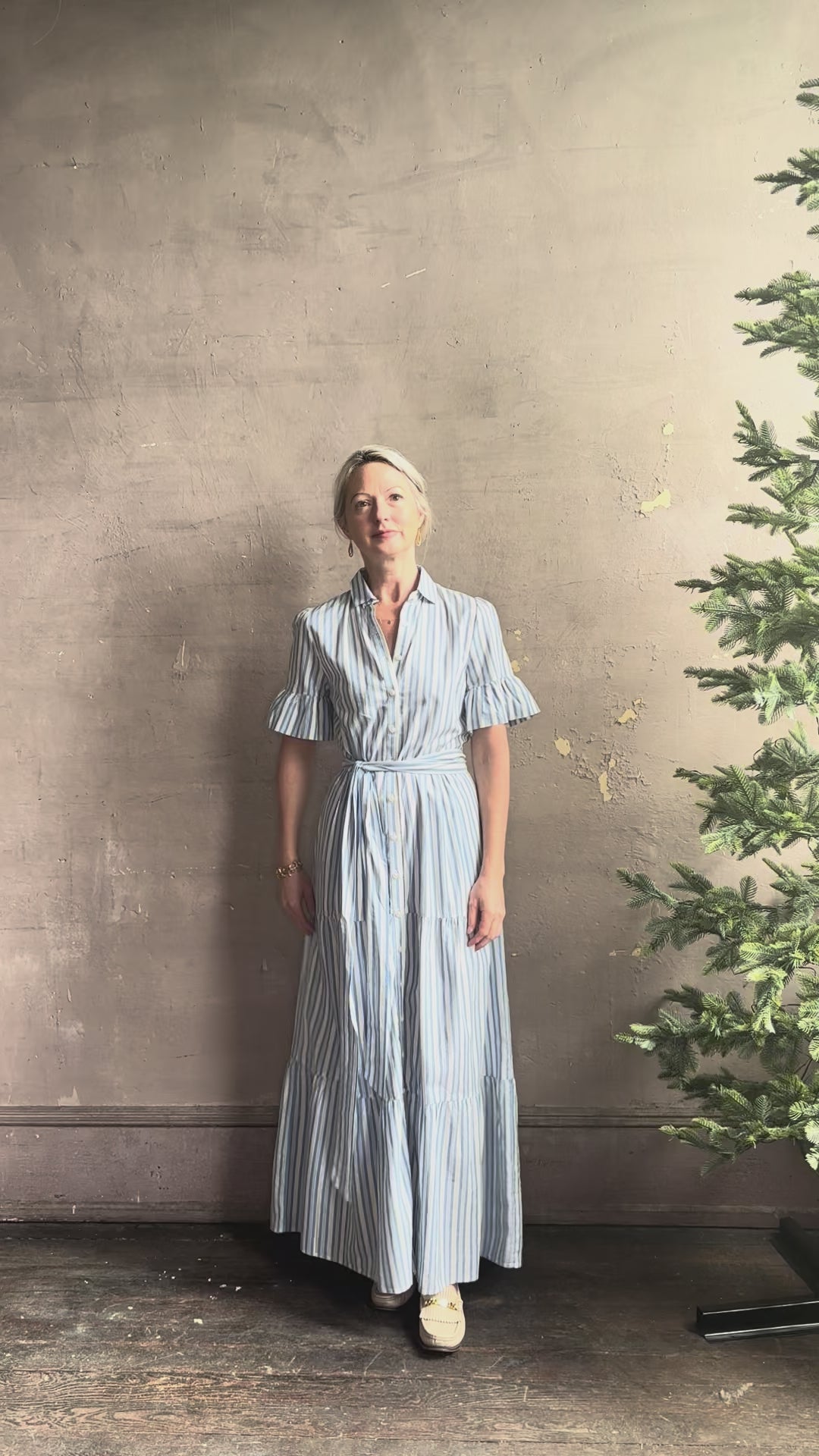 Mature woman reviewing Martine maxi shirt Dress from Trovata rendered in a blue and white Bahia Stripe and crafted from cotton poplin with ruffle lined short sleeves, collar, side seam pockets, button front, tiered skirt and optional self tie co-ordinating fabric belt