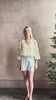 Mature woman wearing Delfina Long Sleeve Silk Blouse from Ulla Johnson rendered in a light yellow body adorned with a delicate, scattered tiny floral print with v-neck, drawstring waist, ruffle lined sleeves and linear ruffle details at front and back