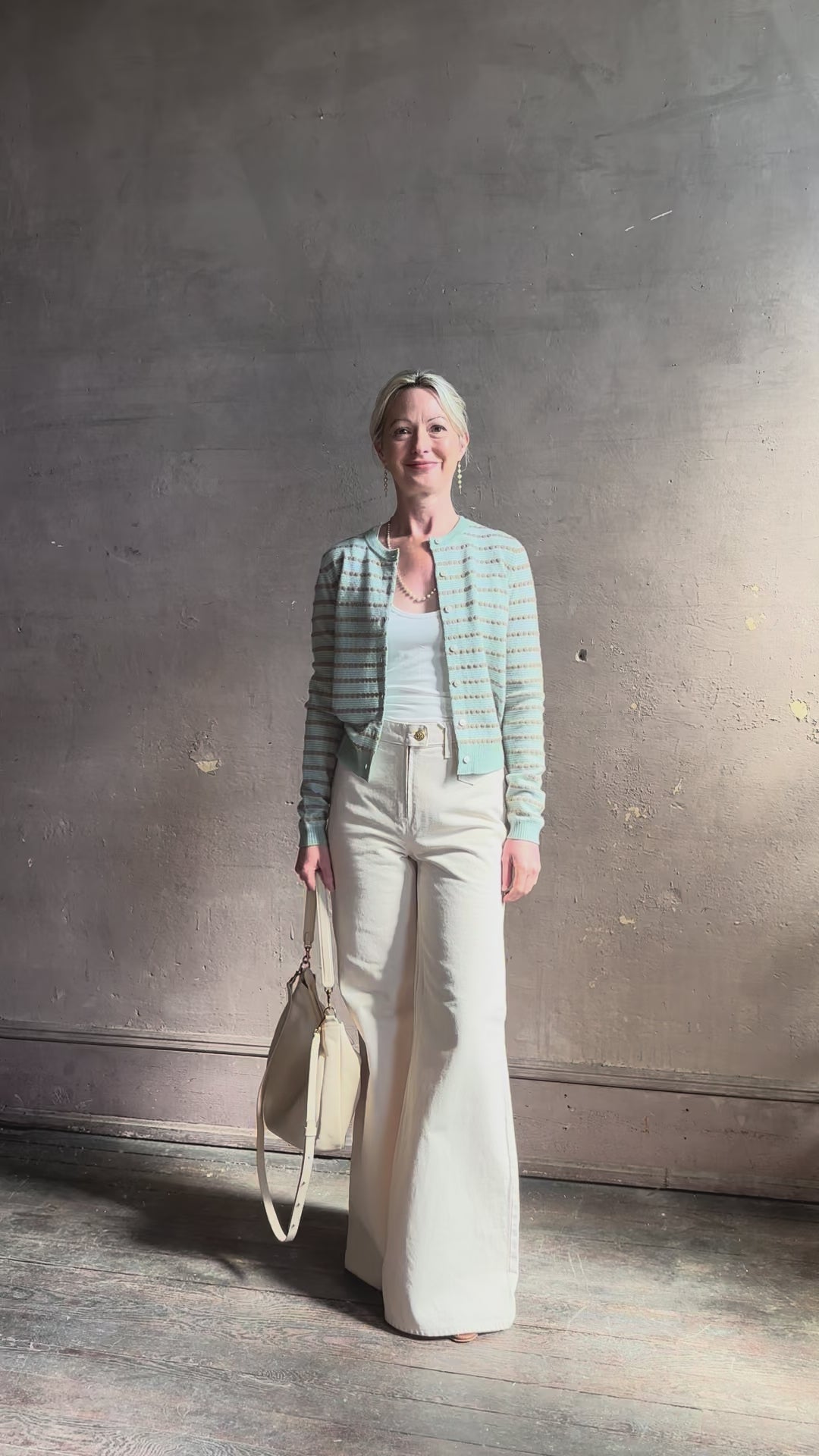 Mature woman reviewing Pebble Stripe Jane Cardigan from Guest in Residence rendered in a serene sage brush aqua Khaki combo and crafted from 100% cashmere with crew neck, long sleeves and button front