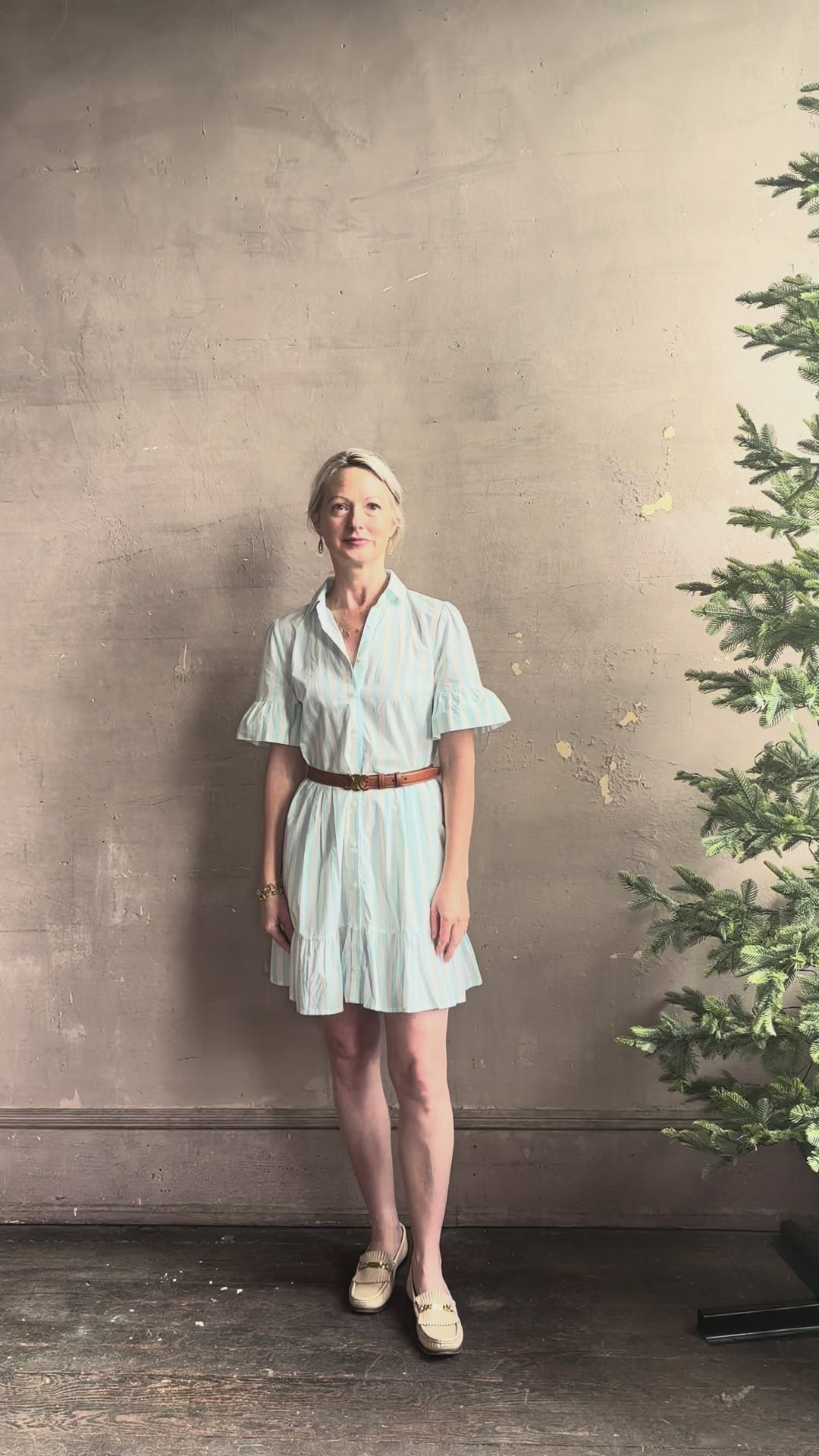 Mature woman reviewing Martinique Dress from Trovata rendered in a cream and aqua Stripe and crafted from cotton poplin with ruffle trimmed short sleeves, collared button front short dress and co-ordinating fabric tie belt