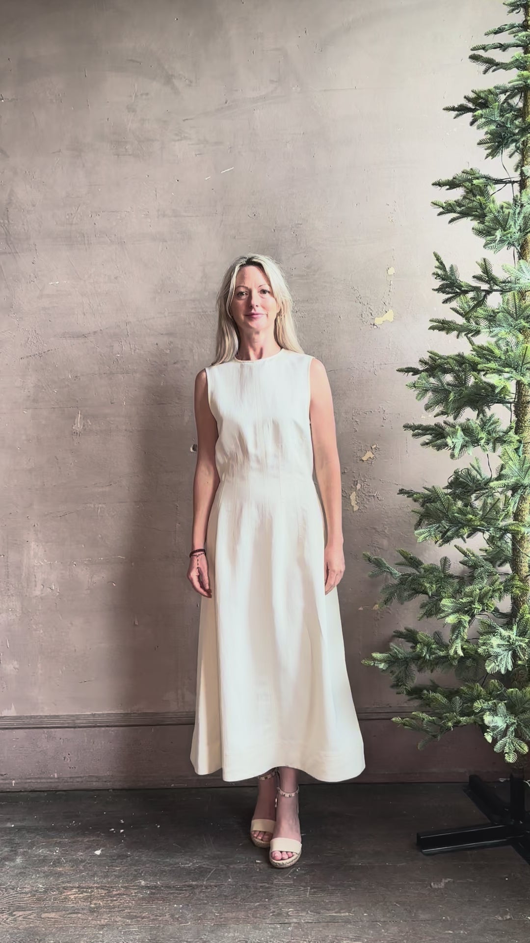 Mature woman reviewing elegant Crew Neck maxi A-Line Dress with Darts from TWP rendered in a soft off white and crafted in the USA from a viscose and linen fabric blend with side seam pockets and sleeveless