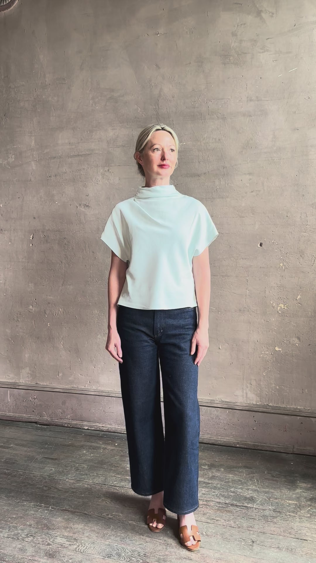 Mature woman reviewing refined Mock Neck Tee Blouse from TWP rendered in a subtle sea glass blue hue and crafted from a viscose linen blend made in the USA, with short sleeves, asymmetrical design and quality fabric draping